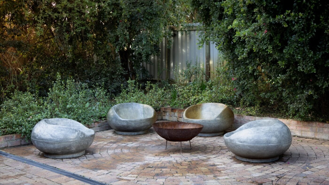 Circular paver patio laid in rings around a metal fire pit bowl, with four stone lounge chairs and garden planting behind