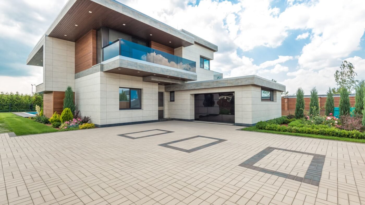 Wide paver driveway in a light running-bond pattern in front of a modern house, the kind of surface finished by compacting pavers into the sand
