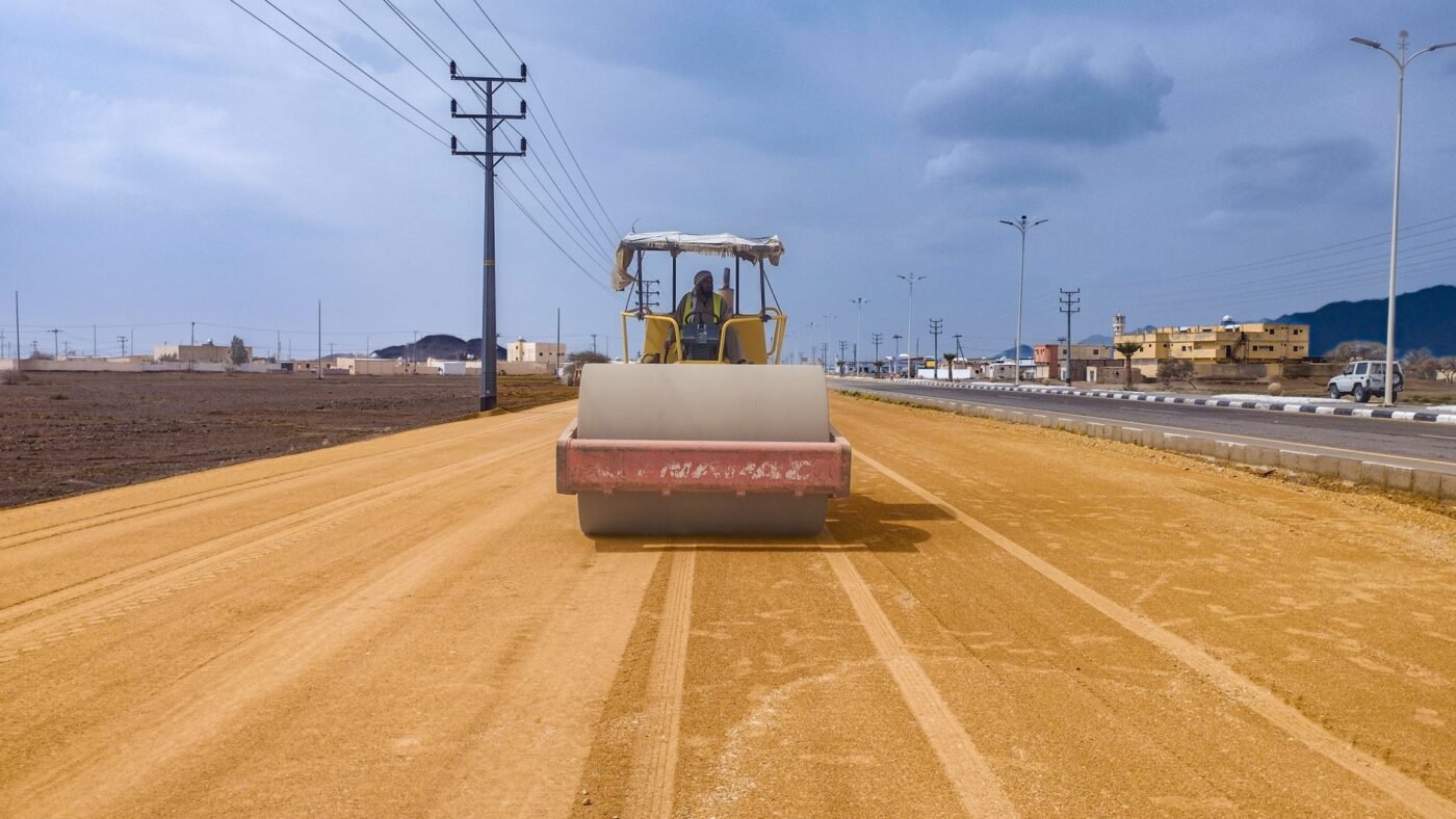Smooth-drum roller compacting a wide strip of graded sandy soil, the kind of subgrade compaction done before a base goes down