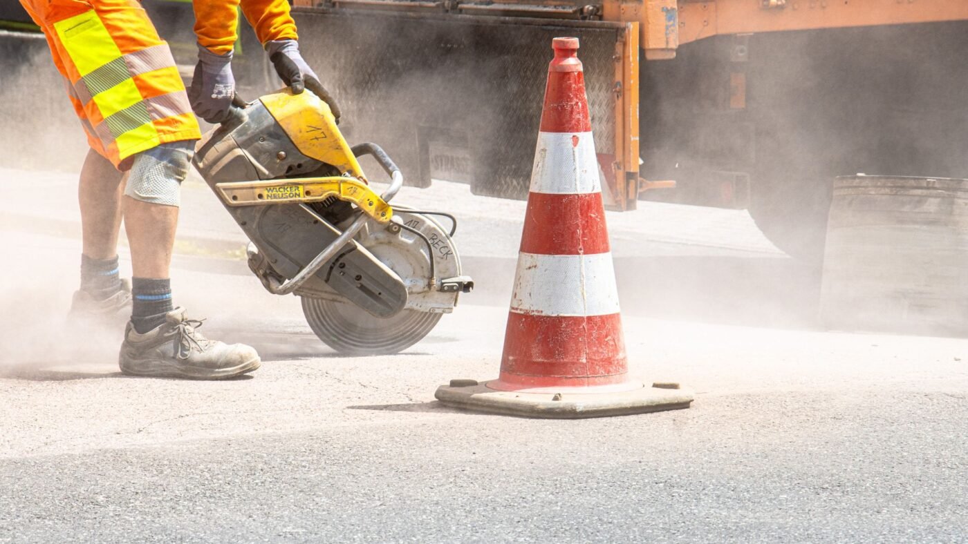 Road worker running a gas cut off saw with a diamond blade across pavement, throwing a cloud of dust
