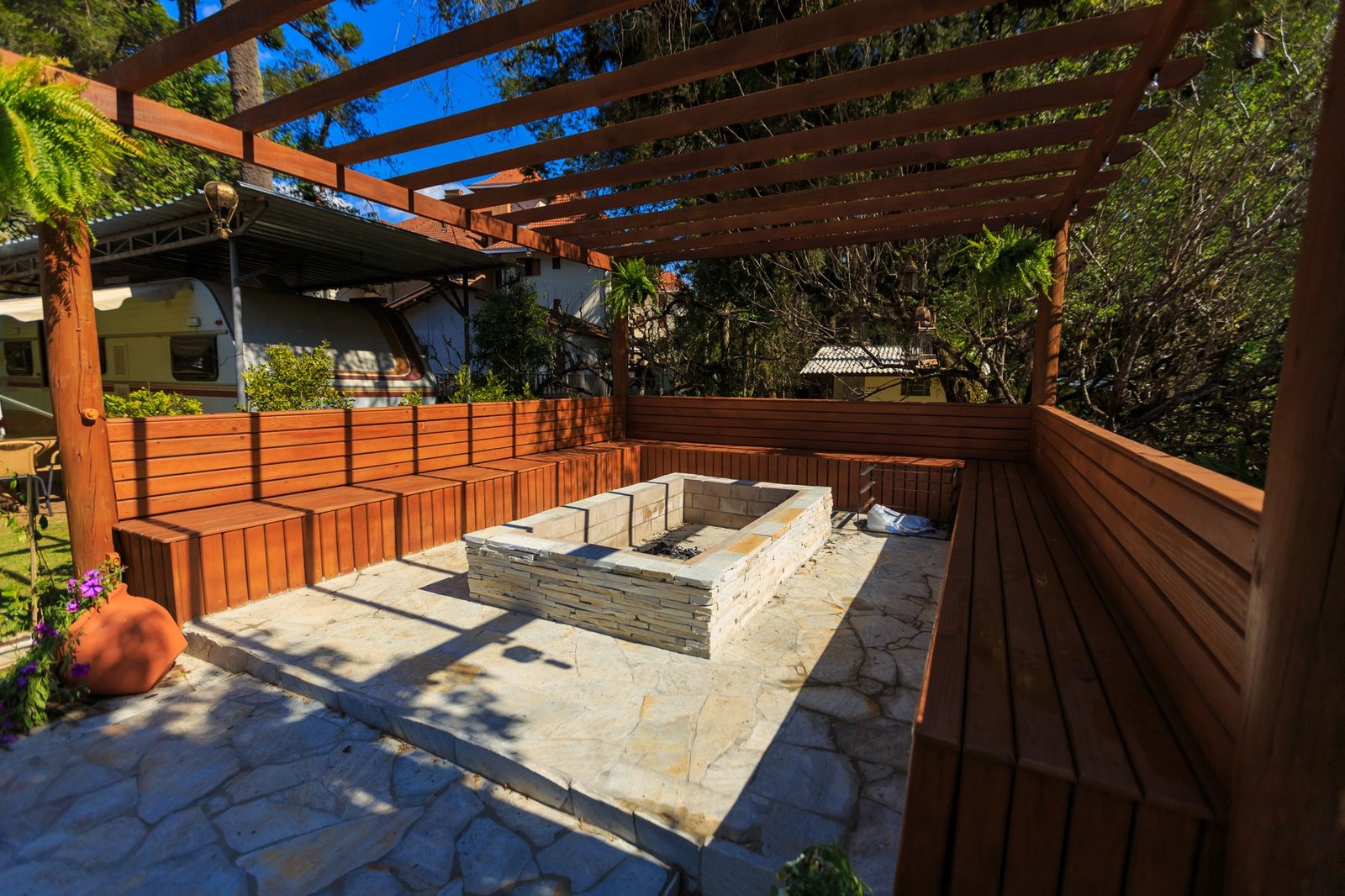 Square stone fire pit set into a flagstone patio under a timber pergola, surrounded by built-in wooden benches