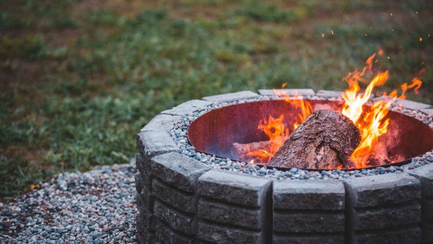 Round fire pit built from gray concrete wall blocks with a steel fire ring insert, burning logs, set on crushed gravel beside a lawn