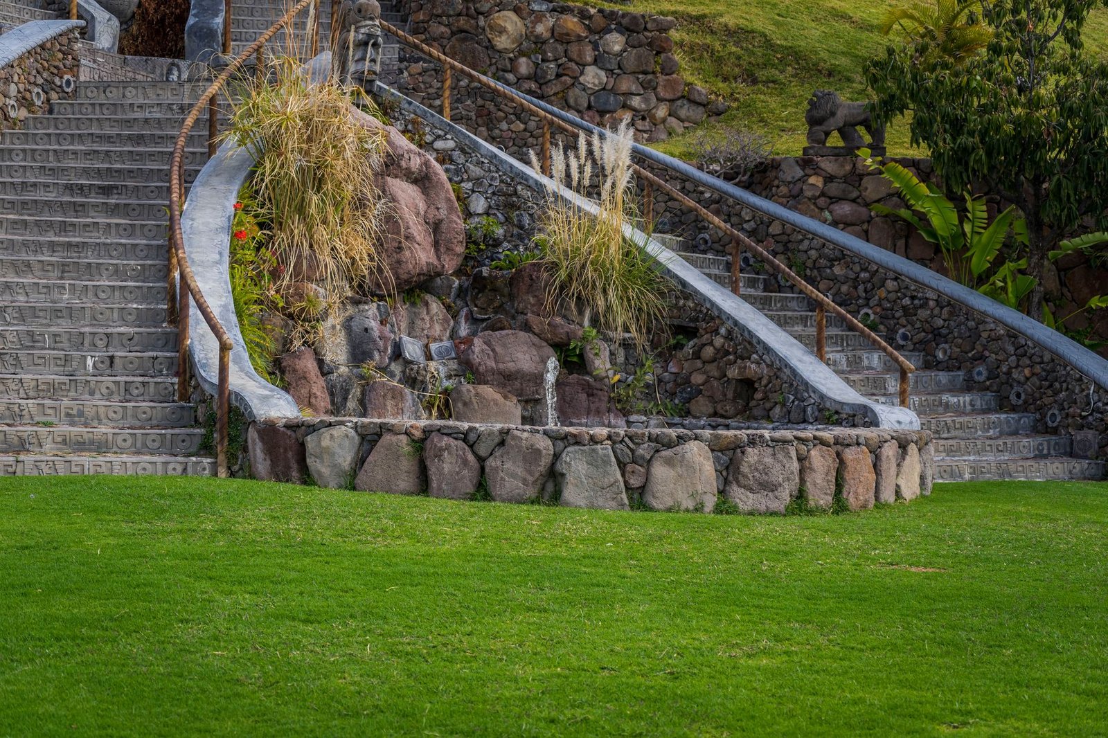 Stone staircase curving up a hillside between stone retaining walls, with ornamental grasses and a green lawn below