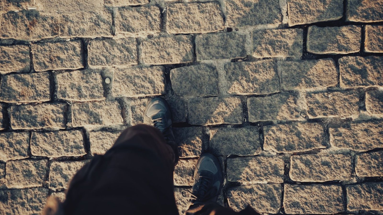 Person's feet in sneakers walking across a sunlit grey cobblestone paver surface
