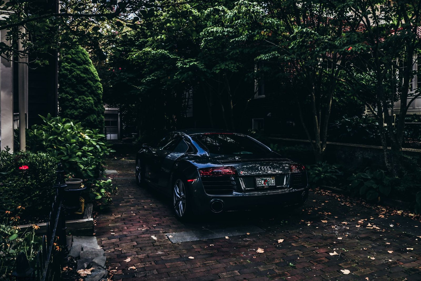 Black sports car parked on a red brick paver driveway under trees, wet leaves on the pavers