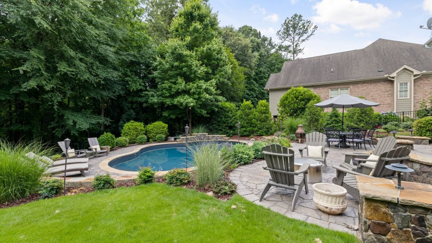 Backyard pool surrounded by a stone paver patio with lounge chairs and a dining umbrella, a sun- and water-exposed surface that needs resealing more often