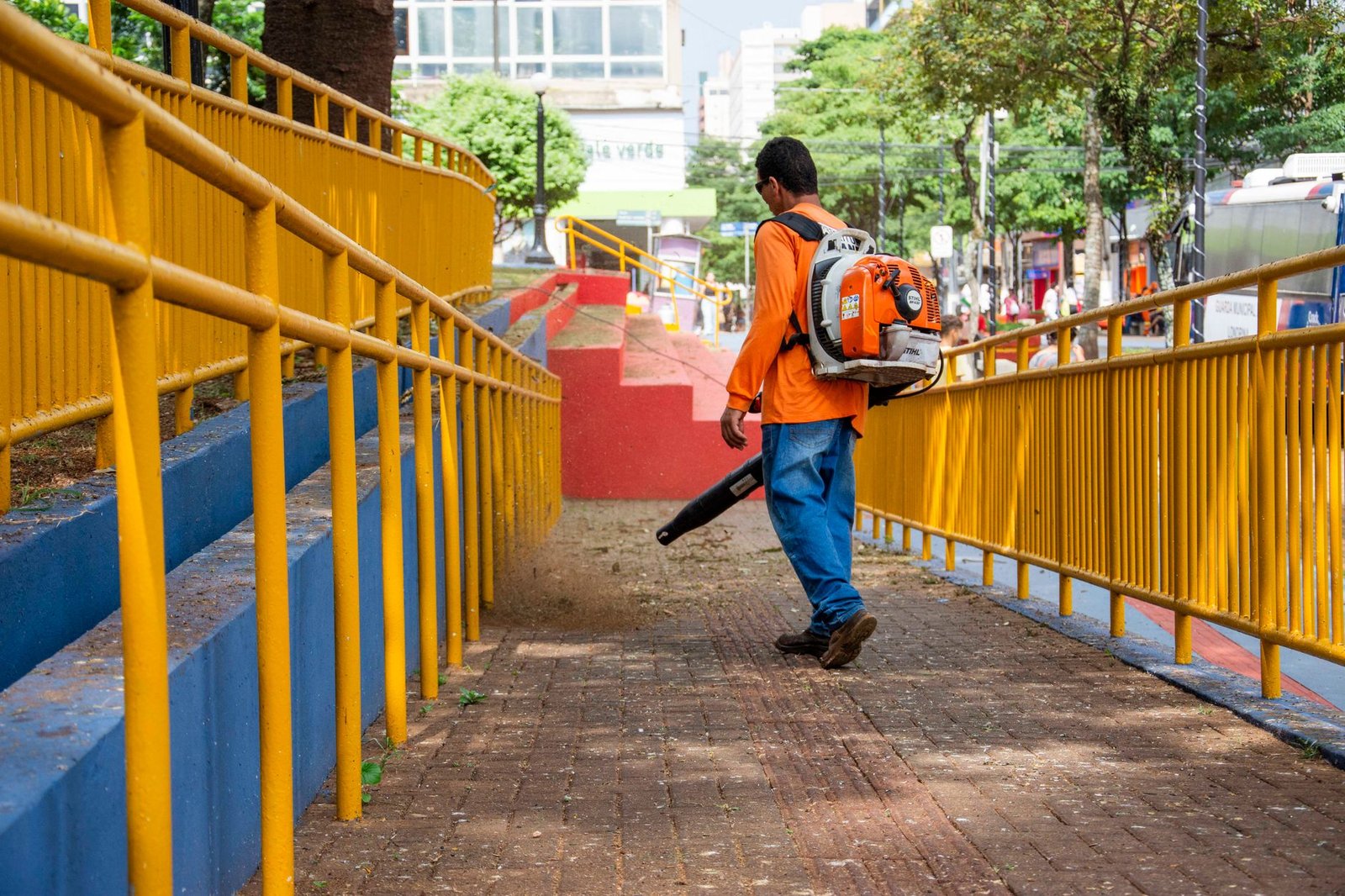 Worker in an orange shirt using a backpack leaf blower on a red brick pavement walkway