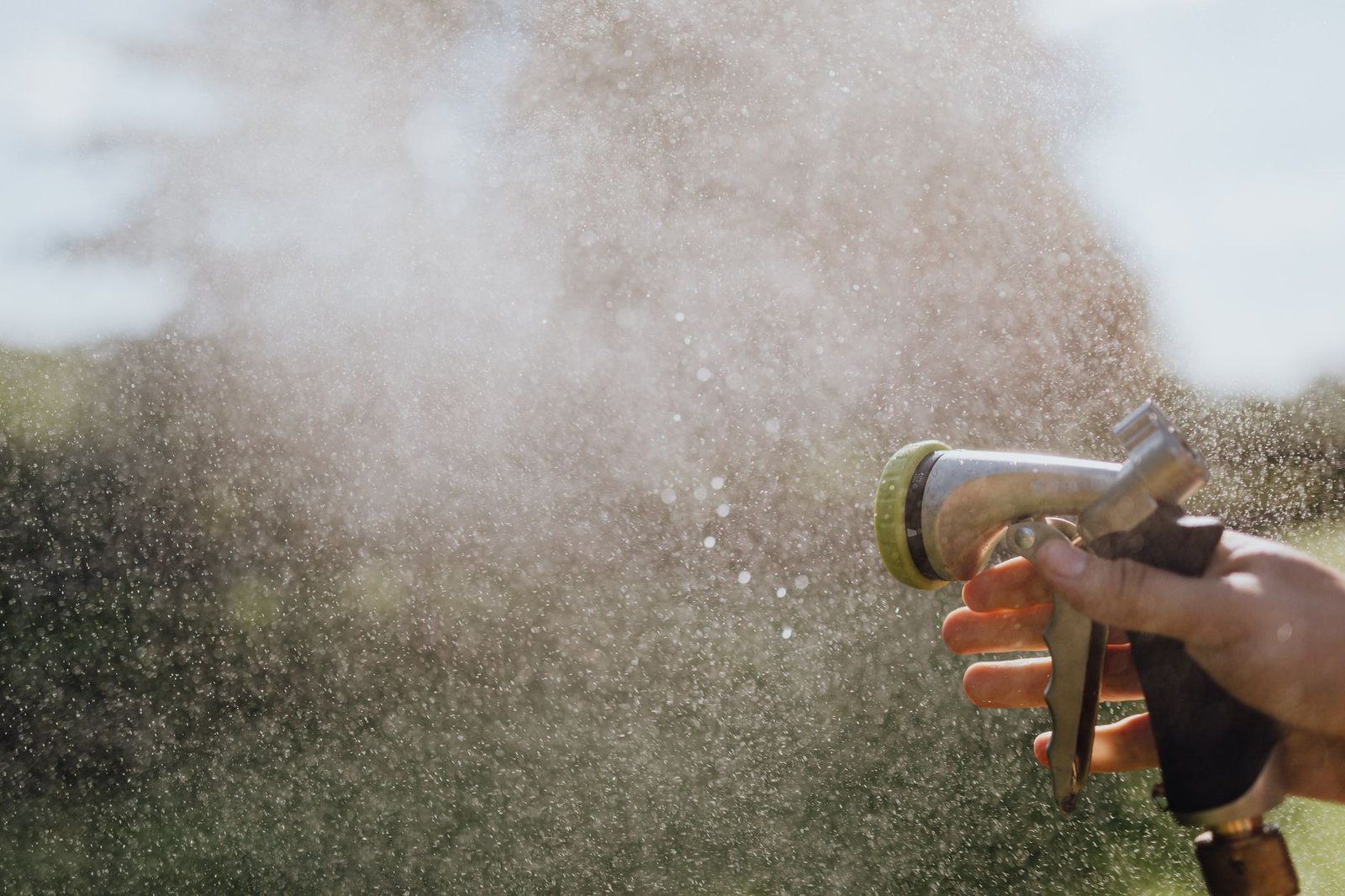 Hand holding a garden hose spray nozzle releasing a fine mist of water outdoors in sunlight