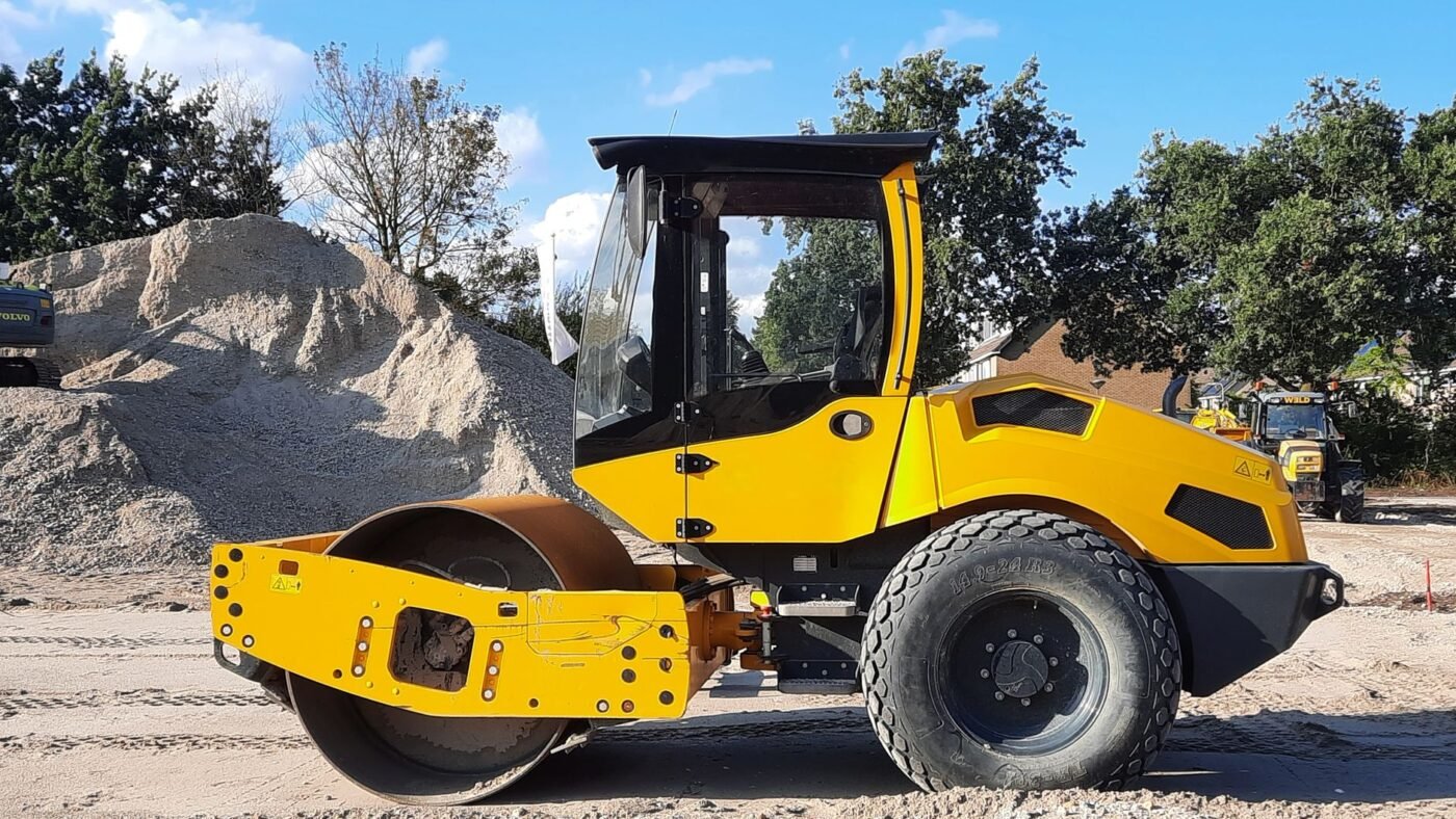Yellow smooth-drum roller parked on a graded gravel base beside a stockpile of crushed stone, ready for compacting gravel