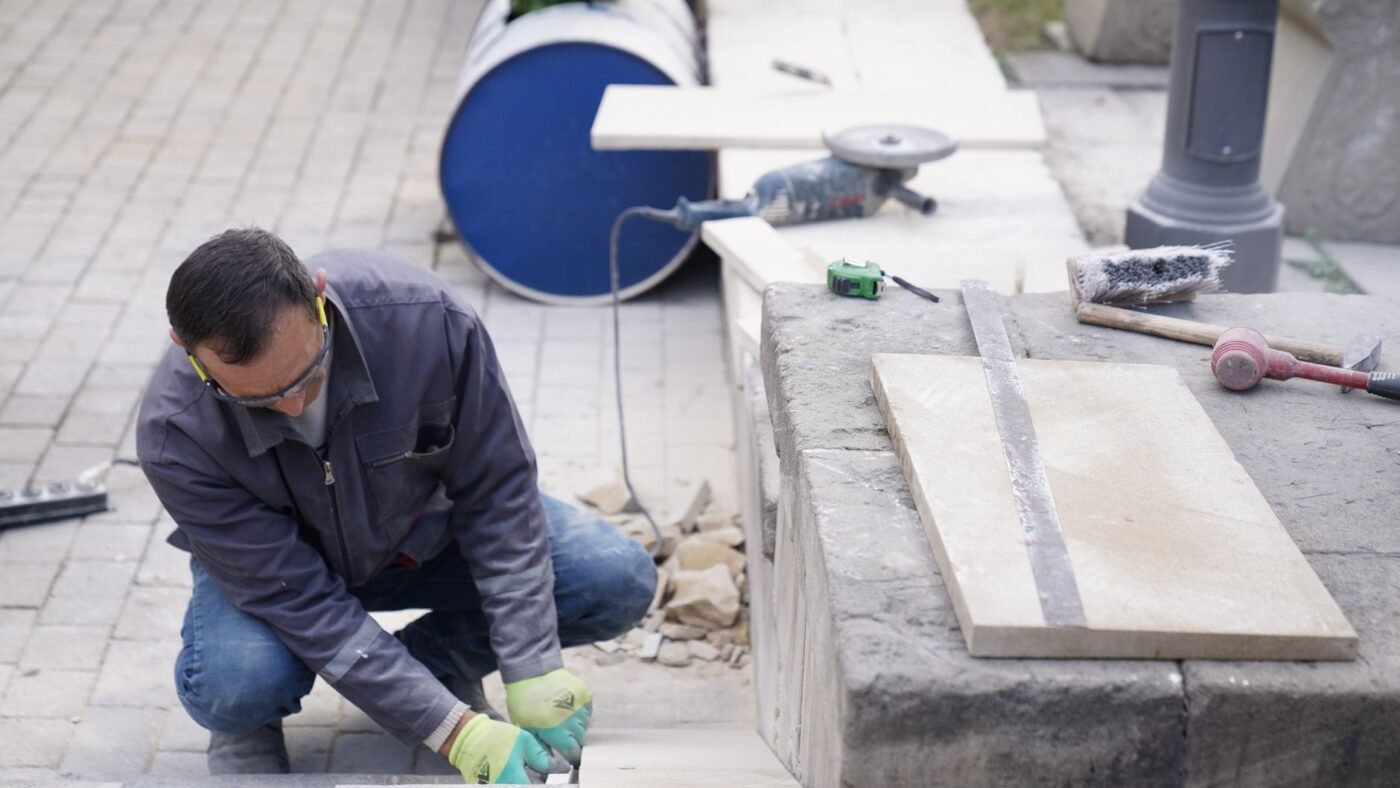 Mason kneeling beside a stone slab marked with a straight cut line, angle grinder and tape measure ready, showing how to cut pavers on site