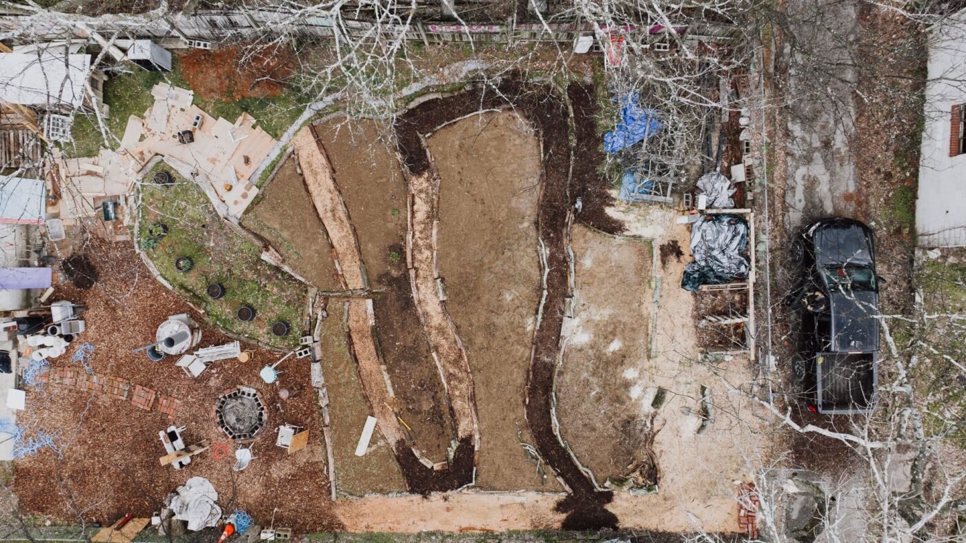 Aerial view of a backyard mid-project with sod stripped and beds and paths dug out, showing ground being shaped and leveled before paving