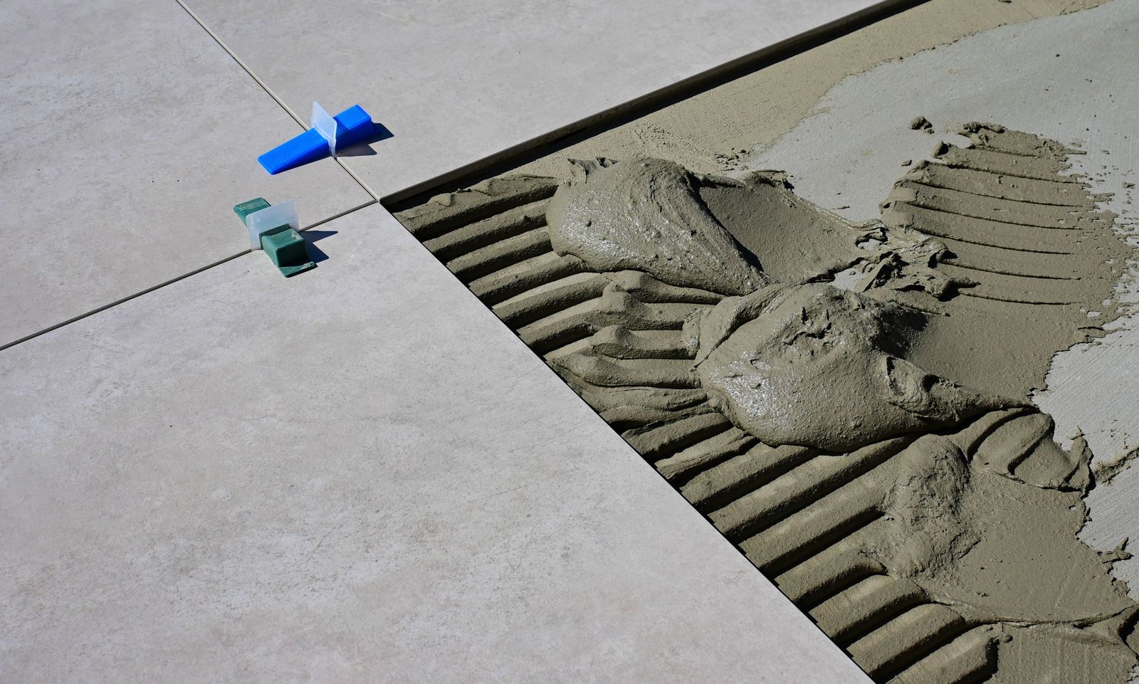 Gloved hand spreading mortar with a notched trowel on a concrete slab beside large-format outdoor pavers held with spacers, setting pavers for mortar joints