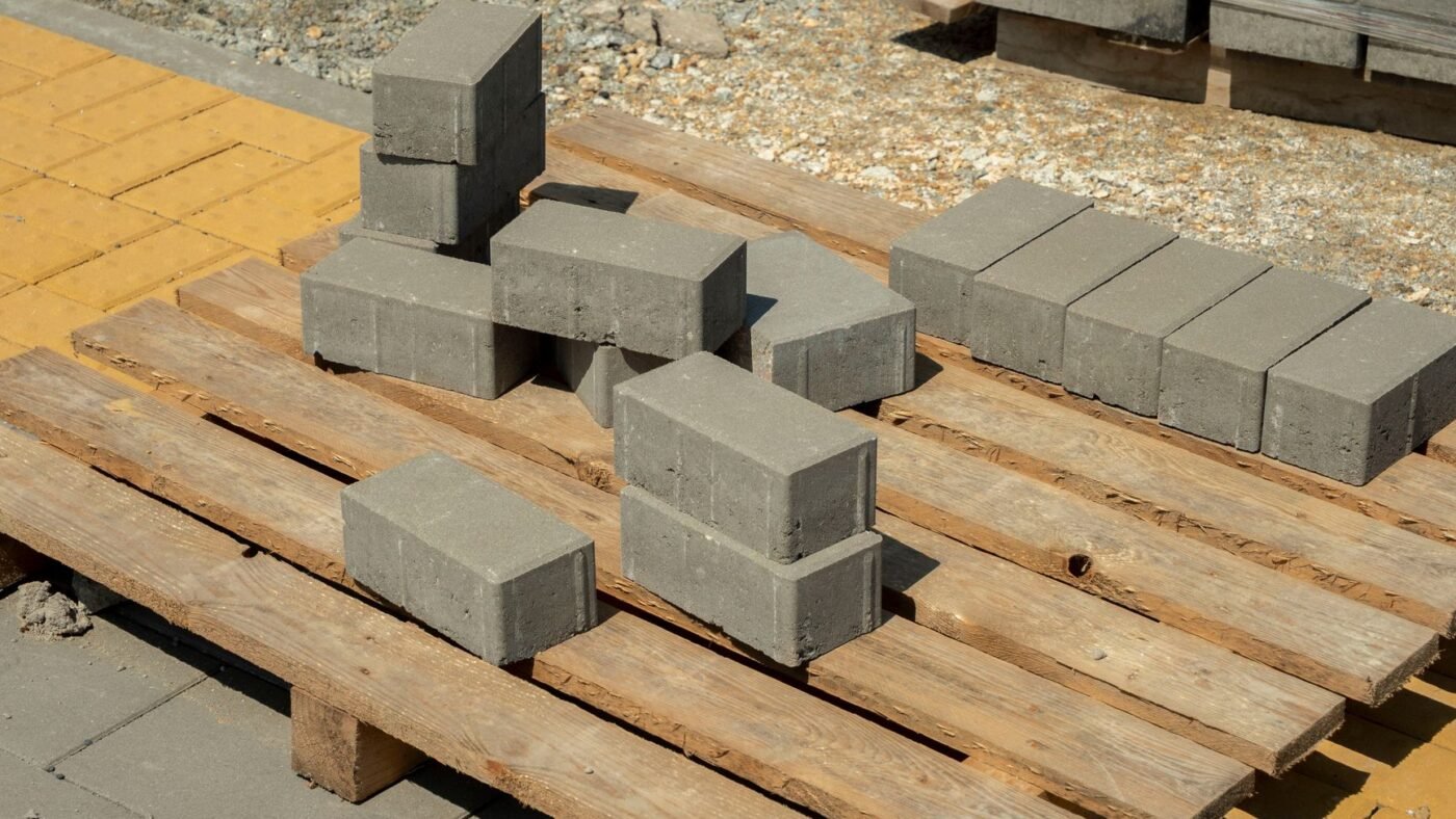Gray concrete pavers stacked on a wooden pallet at a job site, with shrink-wrapped pallets of pavers behind