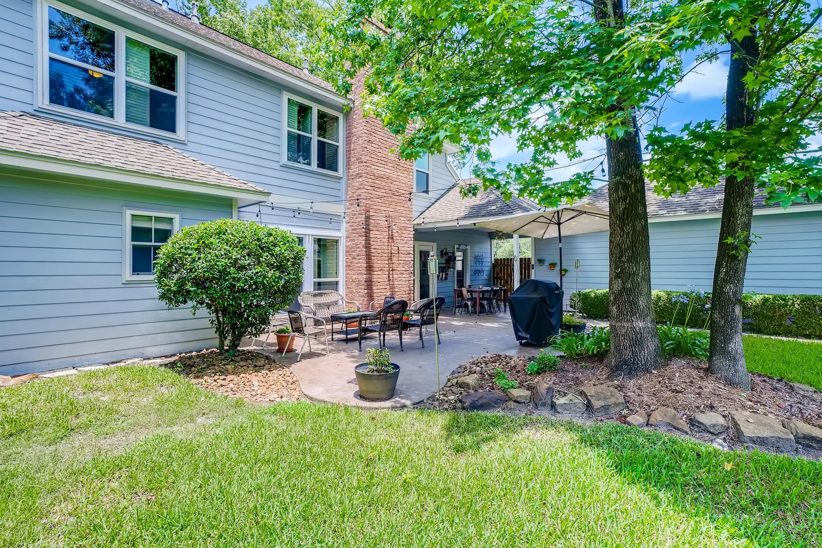 Paver patio beside a house with a planting bed edged by natural stones under a tree