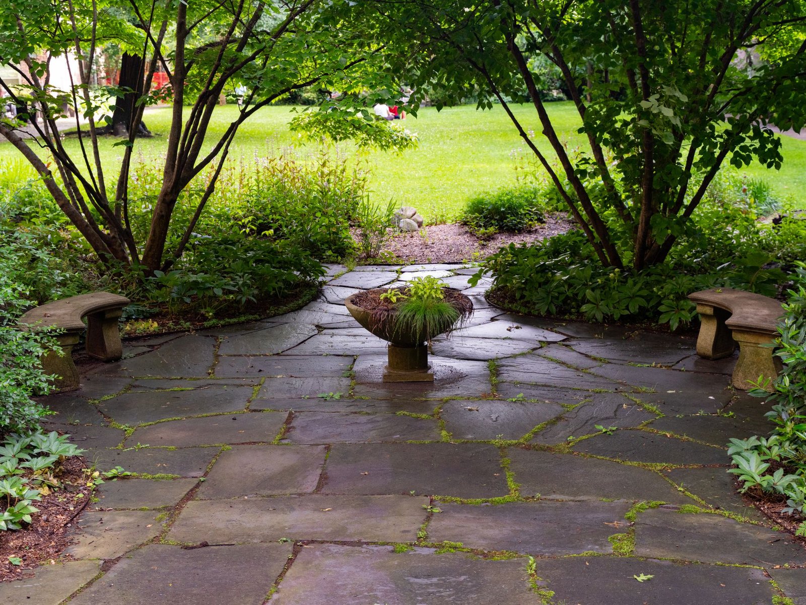 Flagstone patio with stone benches surrounded by planting beds and shrubs, with a lawn beyond
