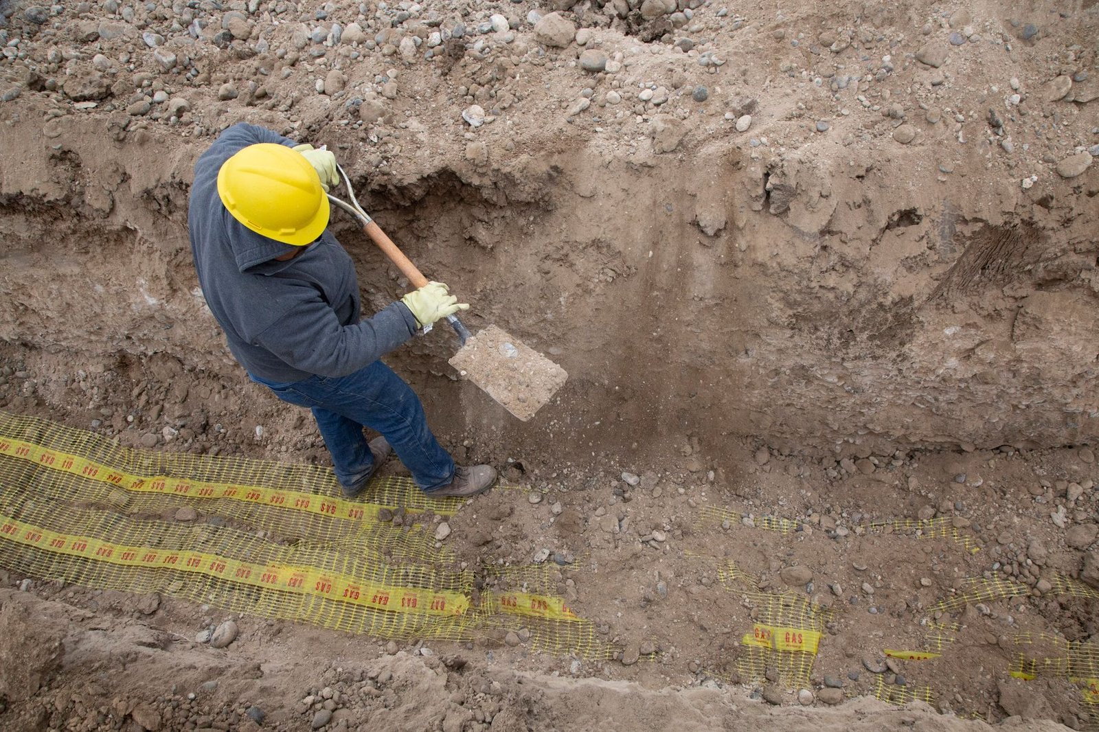 Worker in a hard hat digging an excavation with a shovel to reach firm subsoil below a paver base