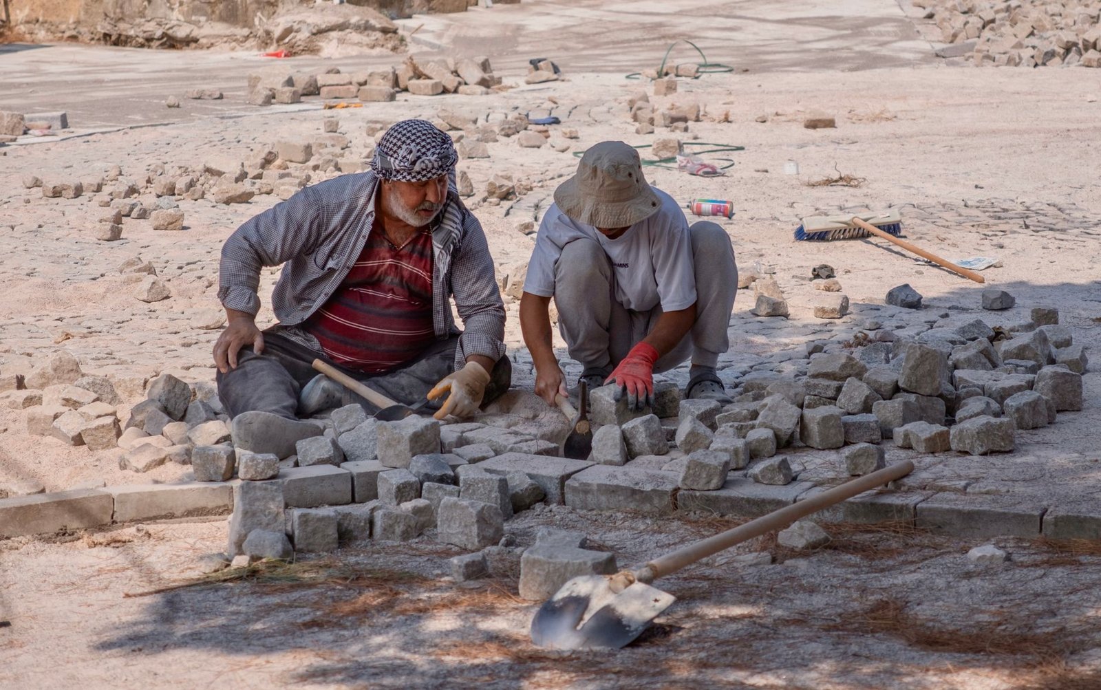 Two workers setting stone pavers by hand against a straight row of rectangular border stones laid first