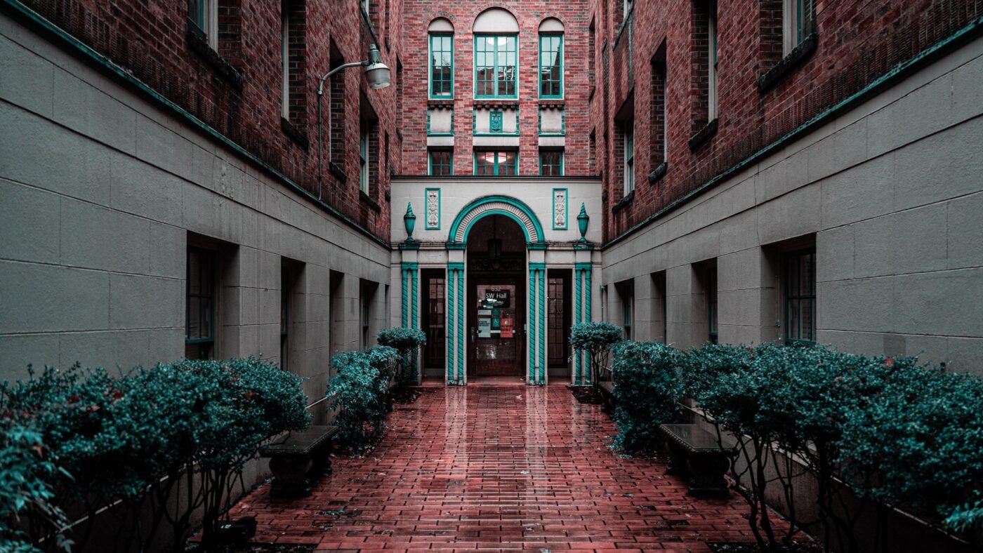 Red paver bricks laid as a walkway through a brick courtyard lined with shrubs, leading to an arched entrance