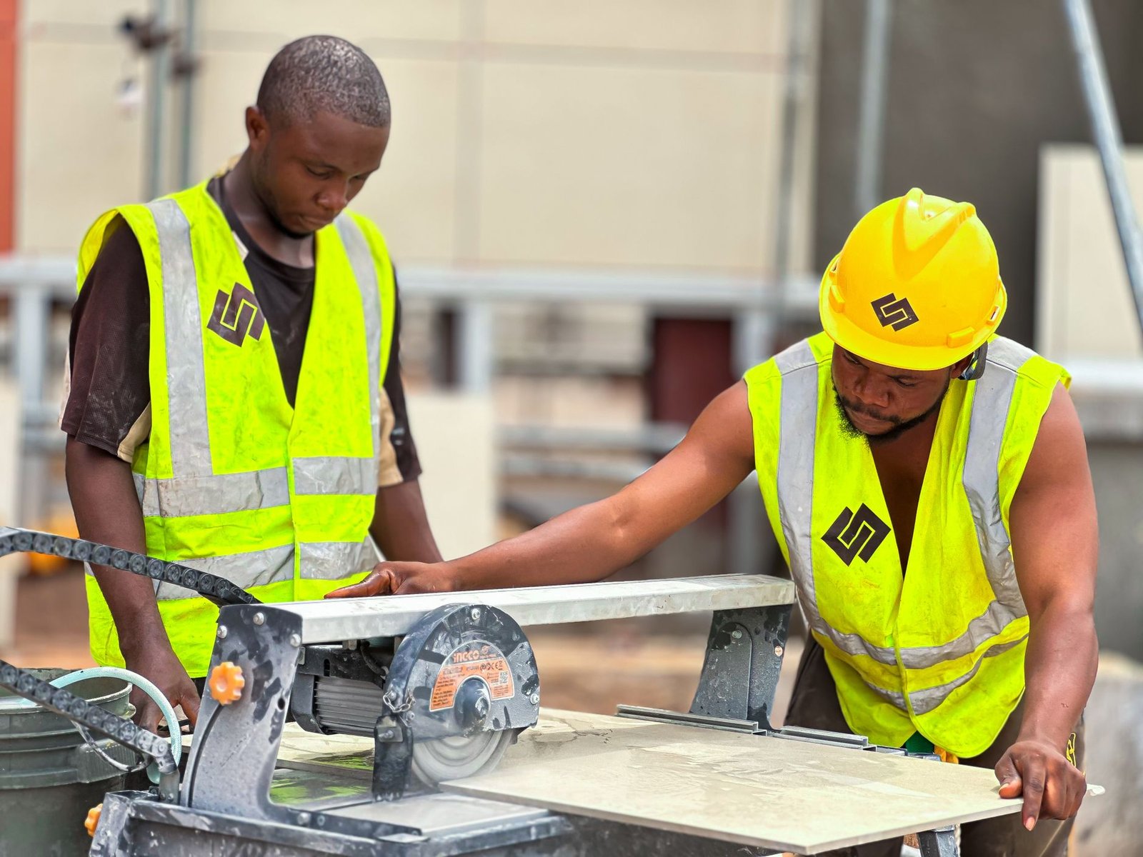 Two workers feeding a large tile across a sliding-table wet saw with a diamond blade, the style of paver cutting machine used for precise cuts