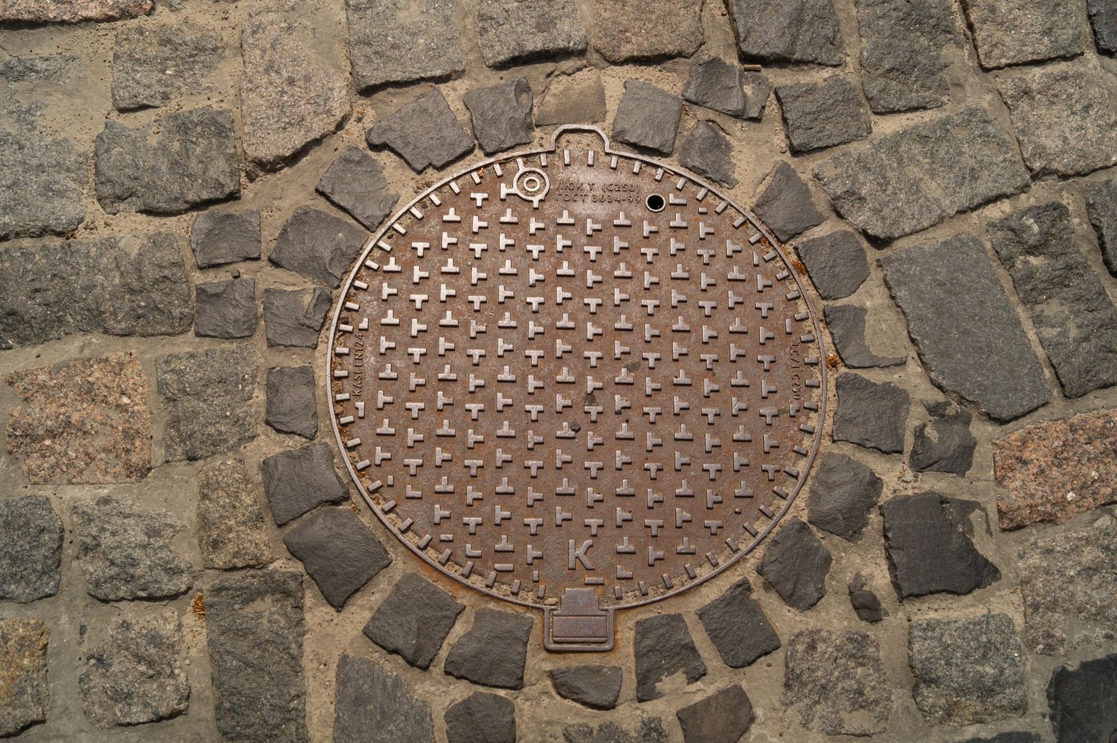 Round cast-iron drain cover set flush into cobblestone paving with the stones cut around its edge