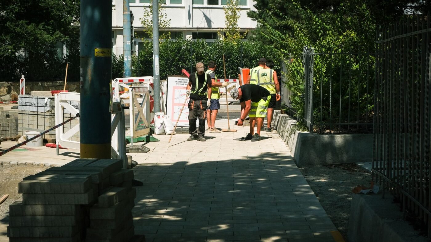 Crew installing a concrete paver sidewalk beside a stack of pavers, with a rigid concrete curb holding the paver edge