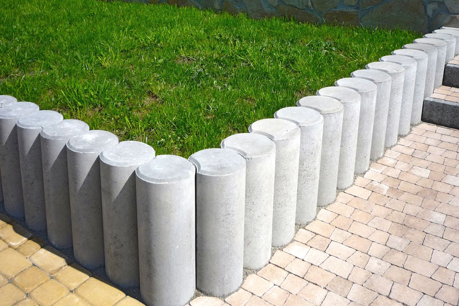 Row of upright round concrete palisade edging stones between a green lawn and a paved area