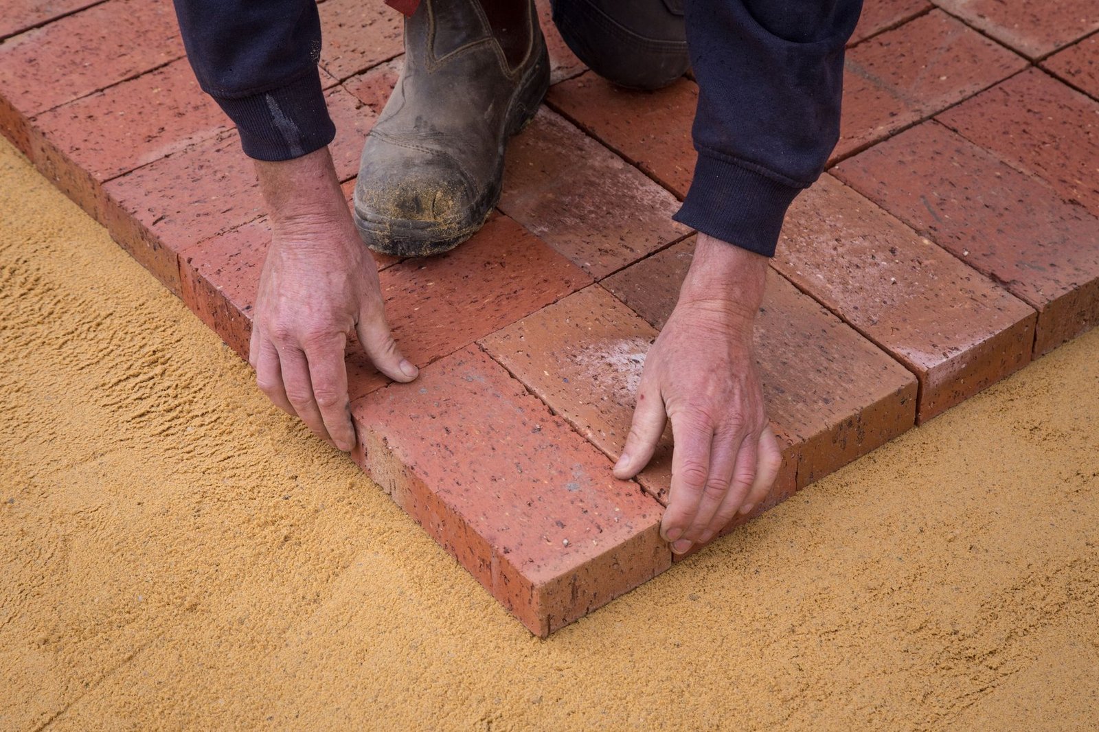 Worker setting red clay pavers onto a smooth screeded sand bed during patio paver installation
