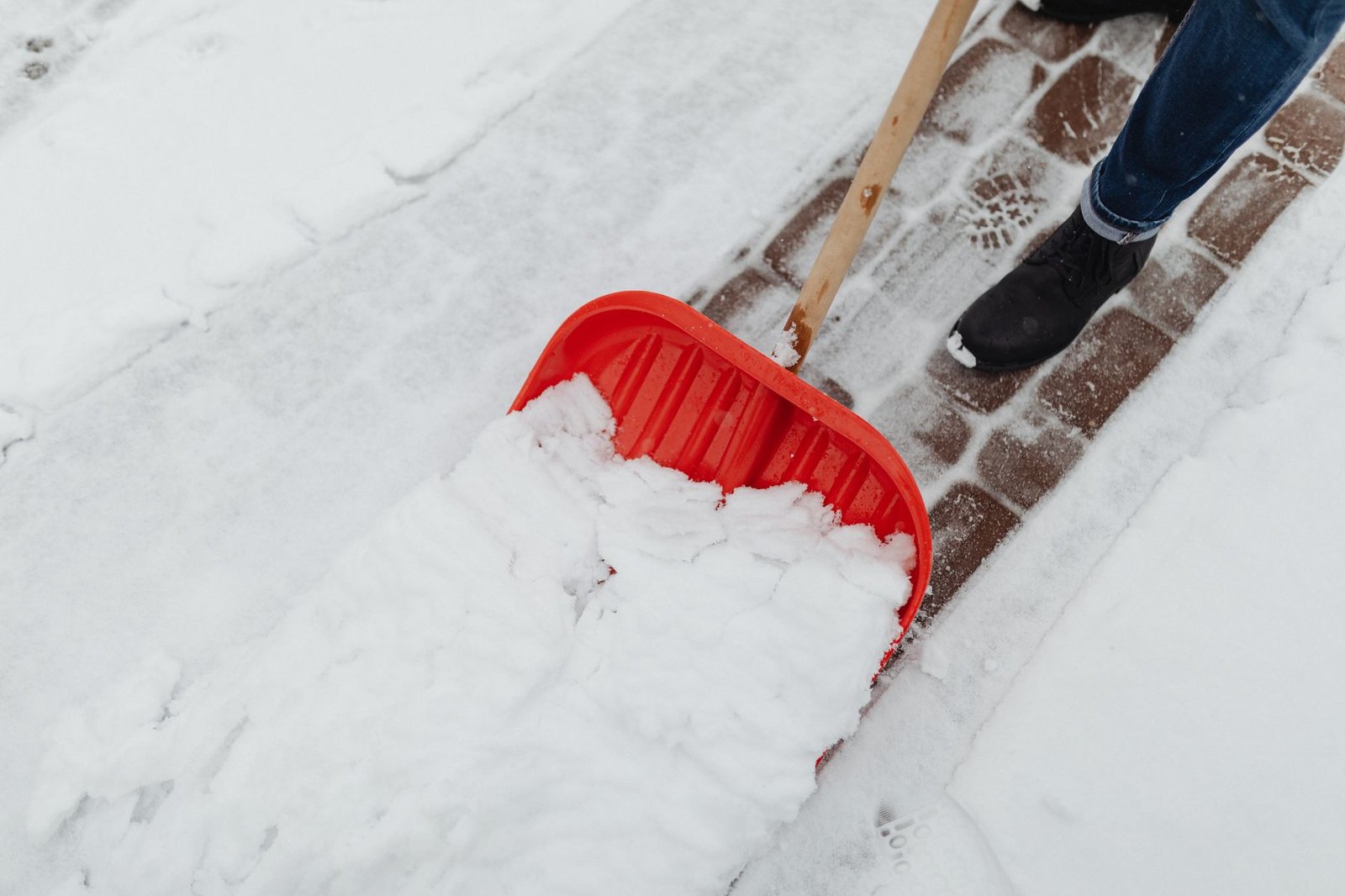 Red plastic snow shovel clearing snow from a brick paver walkway, the right tool for winter paver maintenance