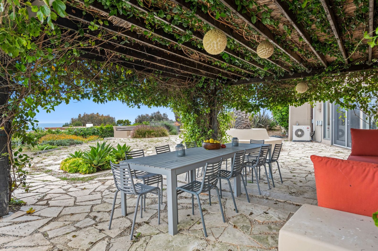 Stone patio under a vine-covered pergola with a long outdoor dining table, string lights and garden beds