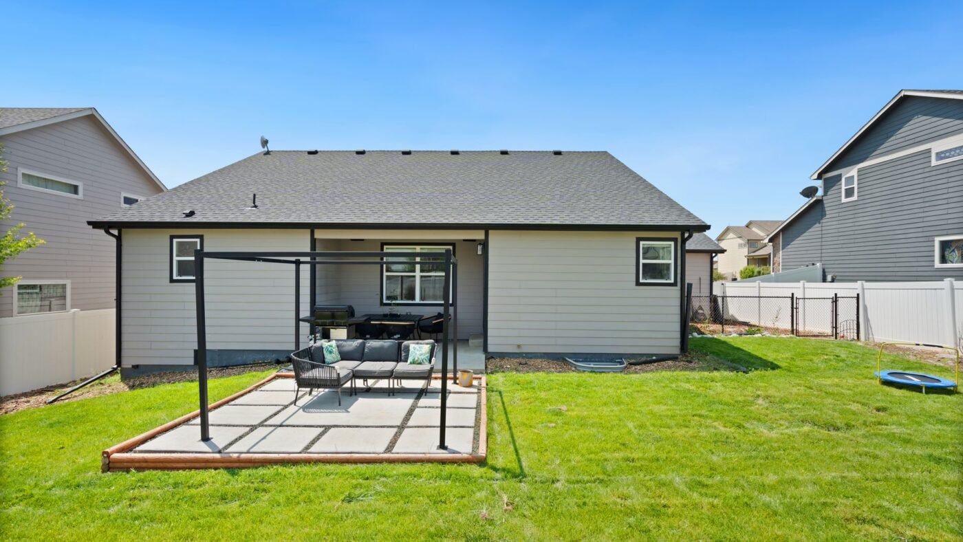 Simple rectangular patio behind a single-story house with an outdoor sofa set and a pergola frame on the lawn