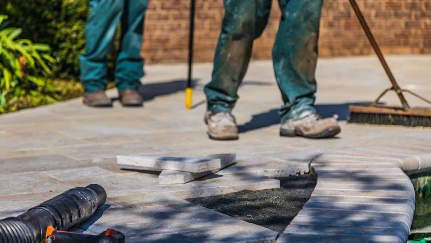 Workers repairing a paver patio with lifted pavers, a push broom and a leaf blower beside the open area