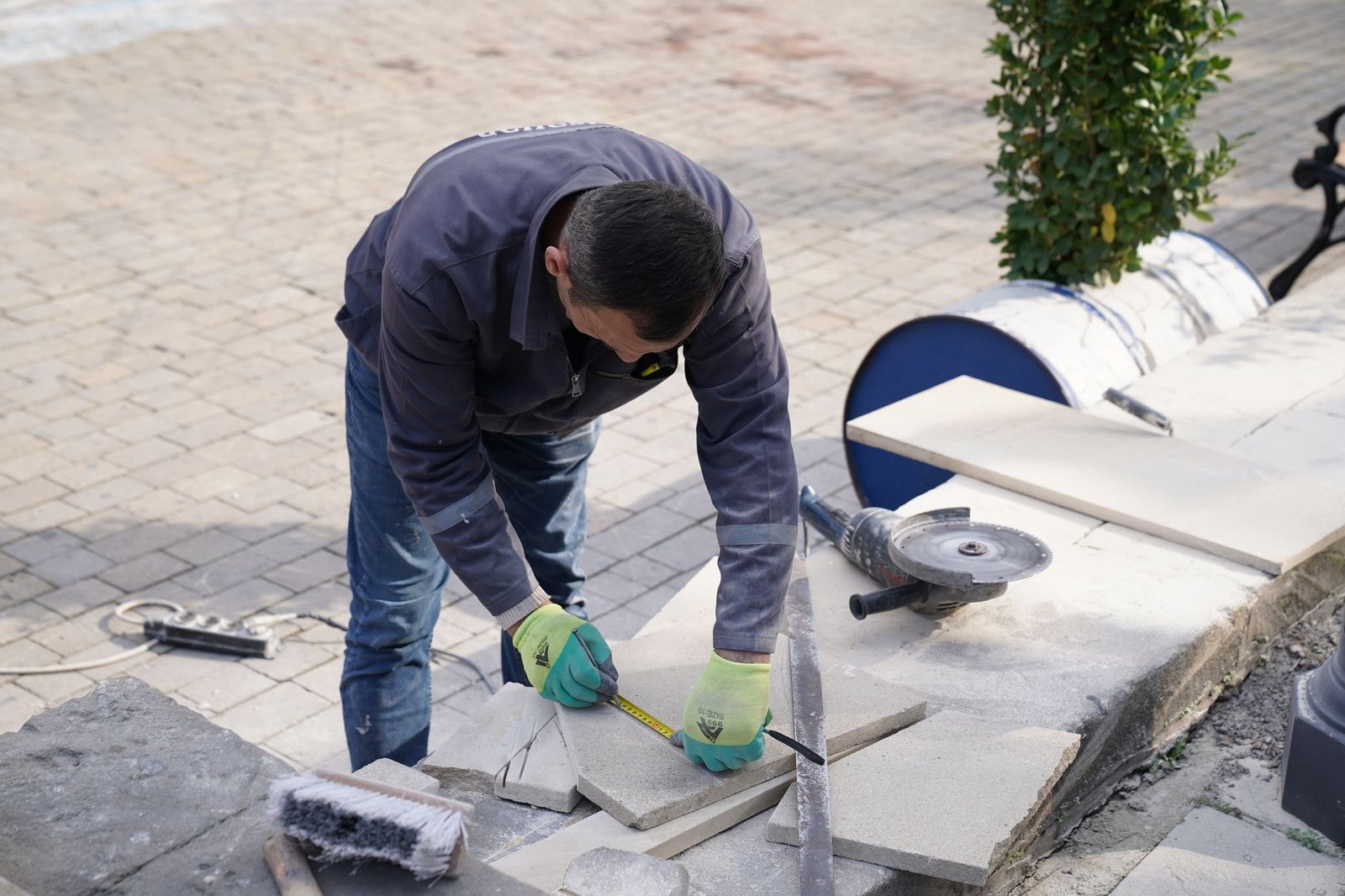 Worker marking a stone slab with a pencil and square beside an angle grinder, preparing a cut on a paving job
