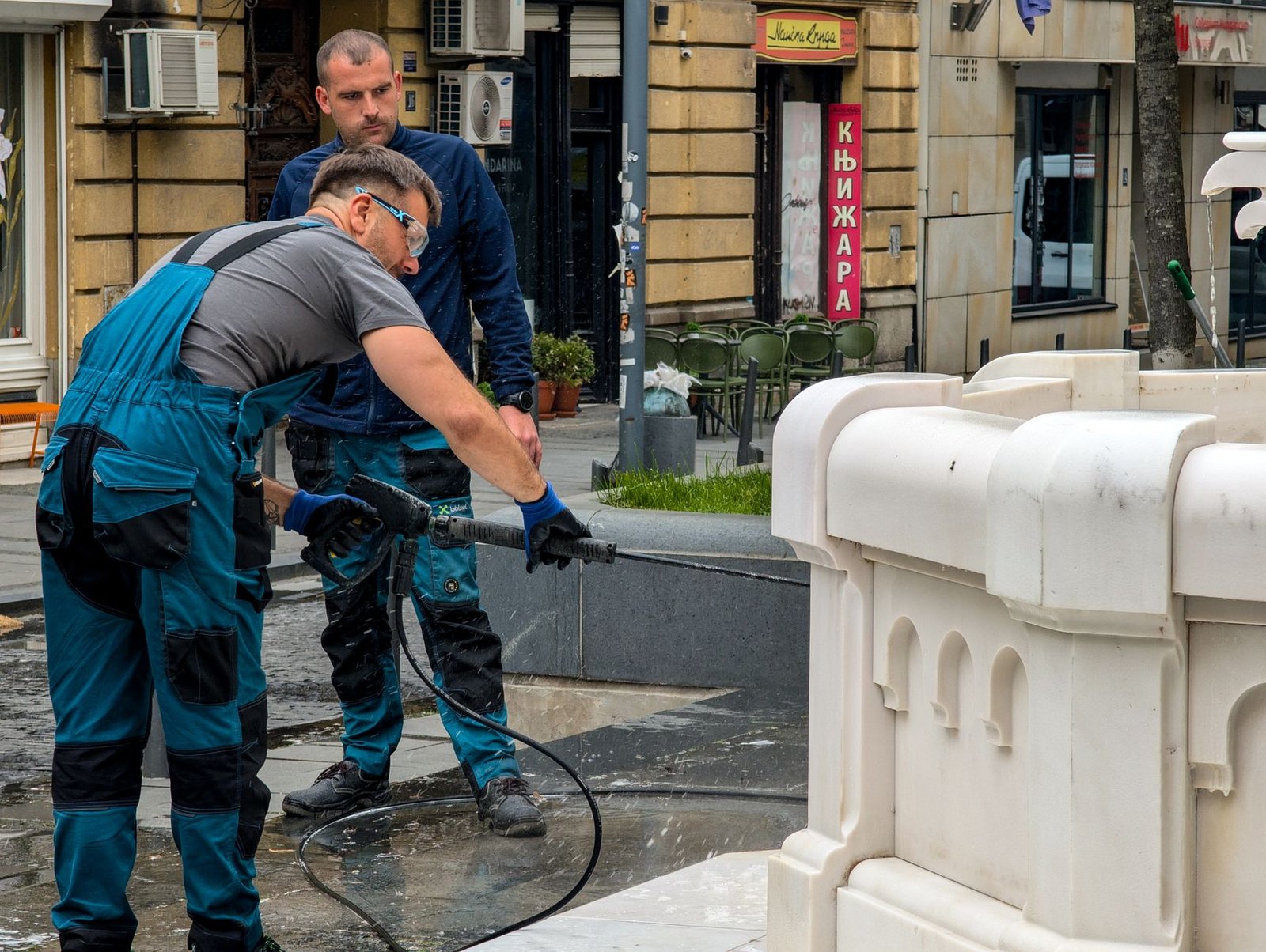 Two workers pressure washing wet stone paving beside a fountain