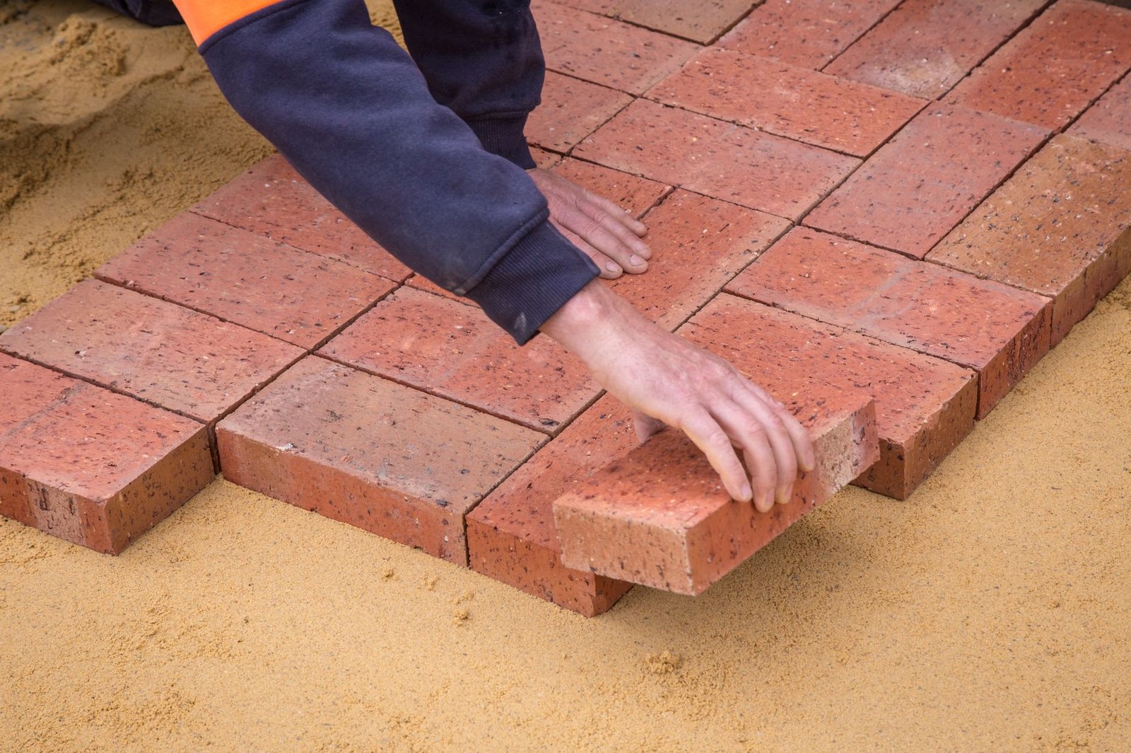 Installer pressing a red paver into place on a screeded sand bed while building a paver walkway