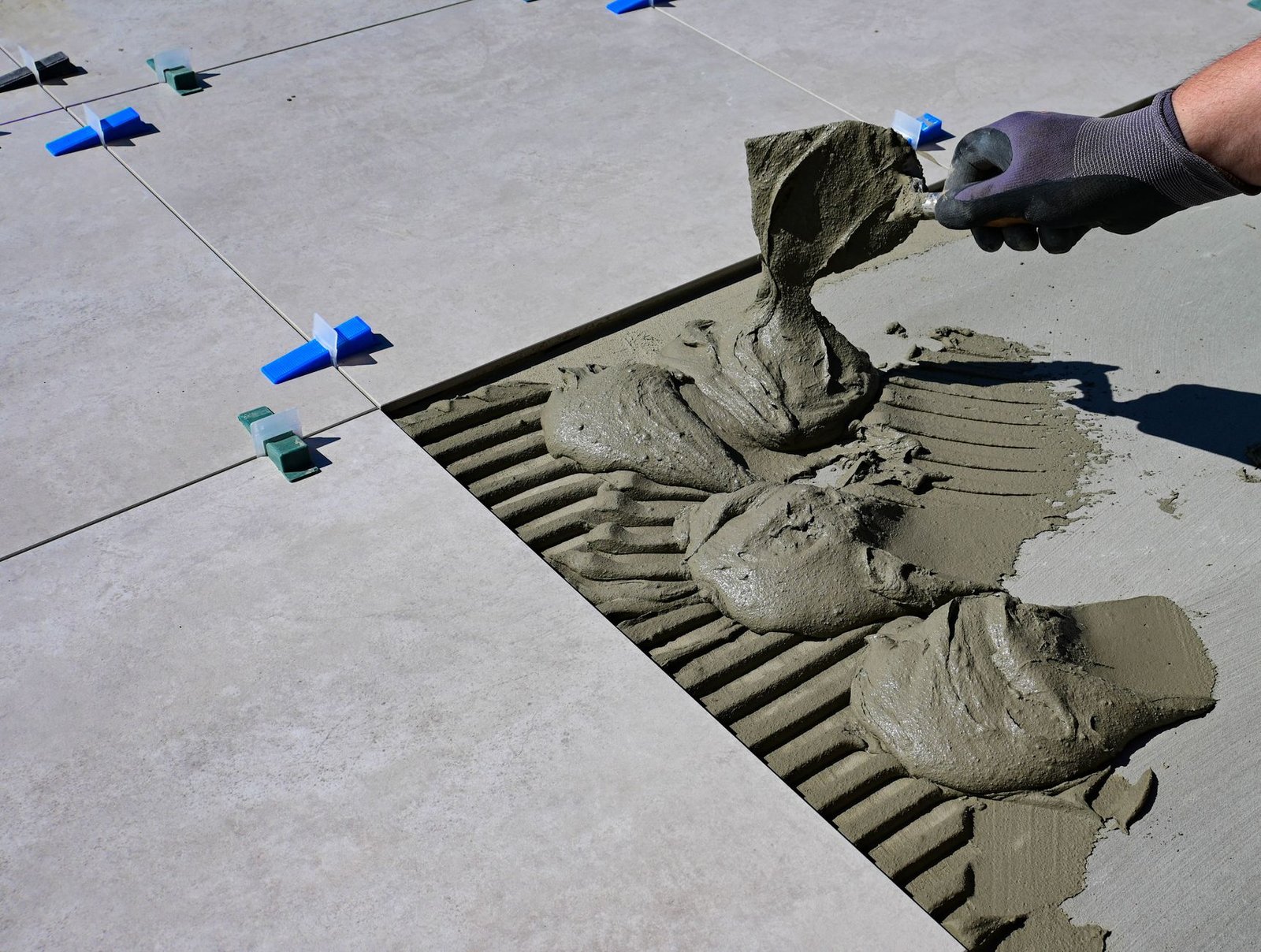 Gloved hand applying mortar with a trowel before setting an outdoor paver, as in a mortar-set installation of pavers on a concrete slab
