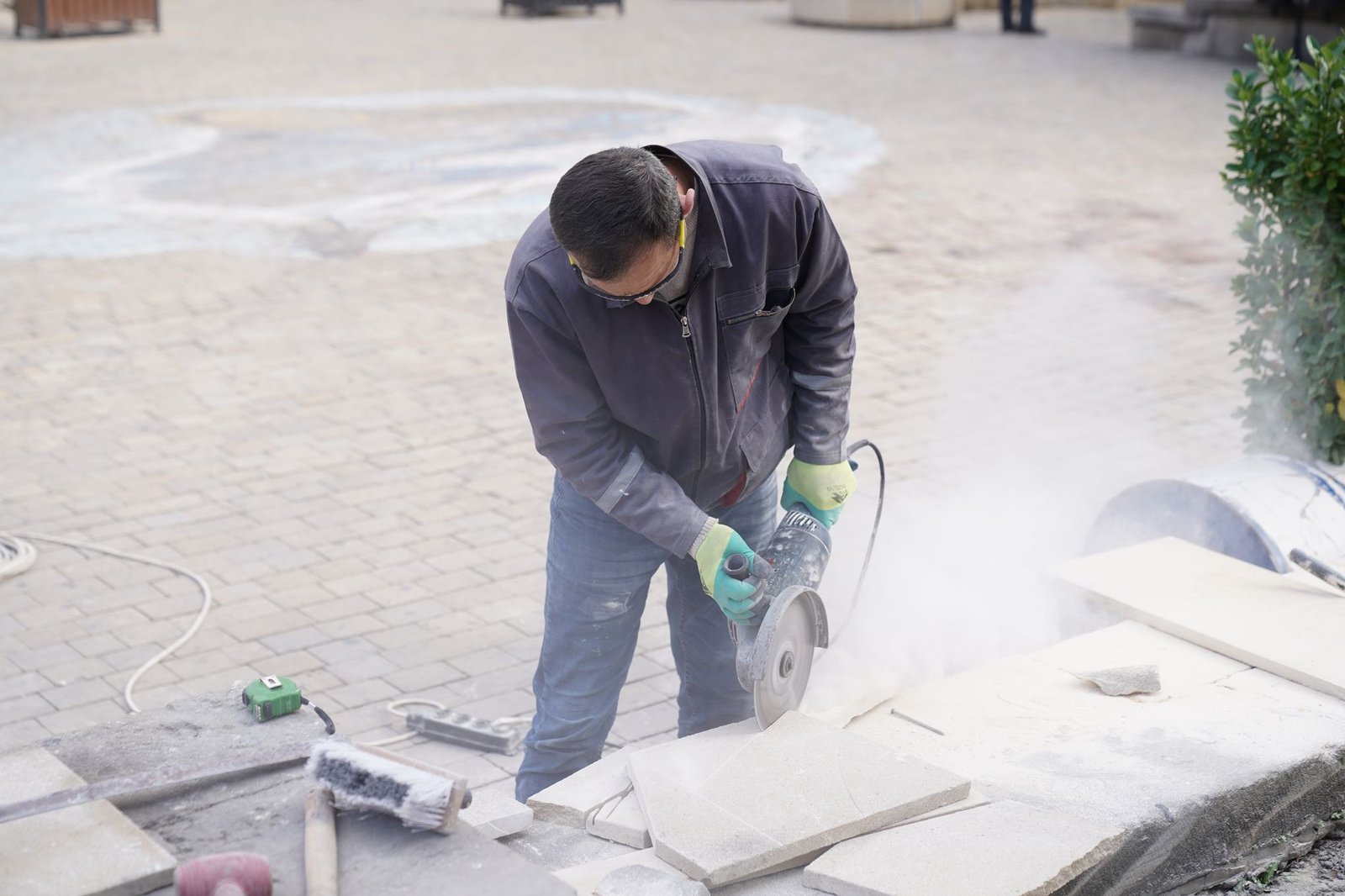 Worker cutting a stone paver with an angle grinder beside a paved area, the kind of cut needed when fitting thin pavers over concrete