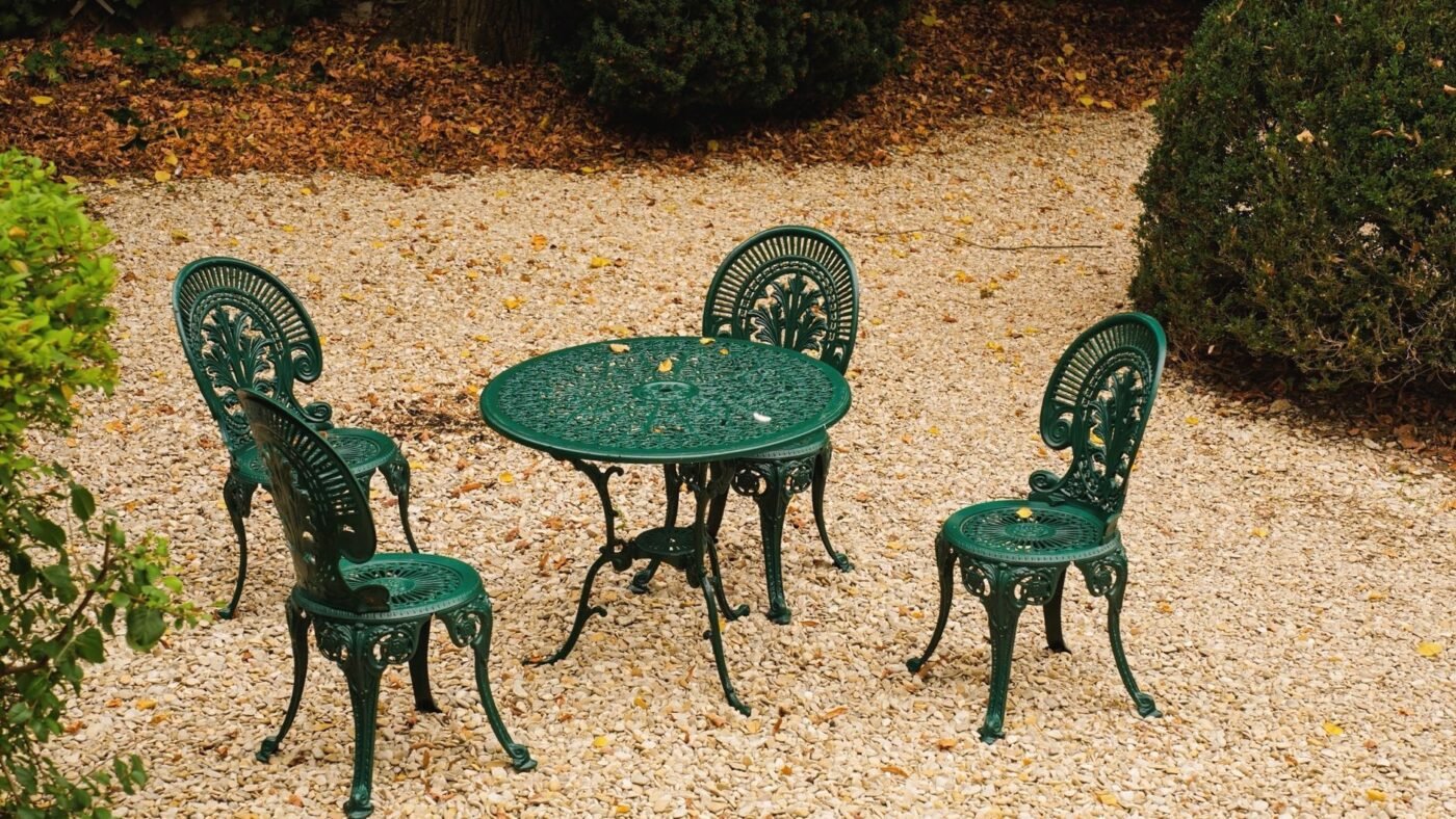 Green cast-metal bistro table and chairs on a pea gravel patio area edged by shrubs