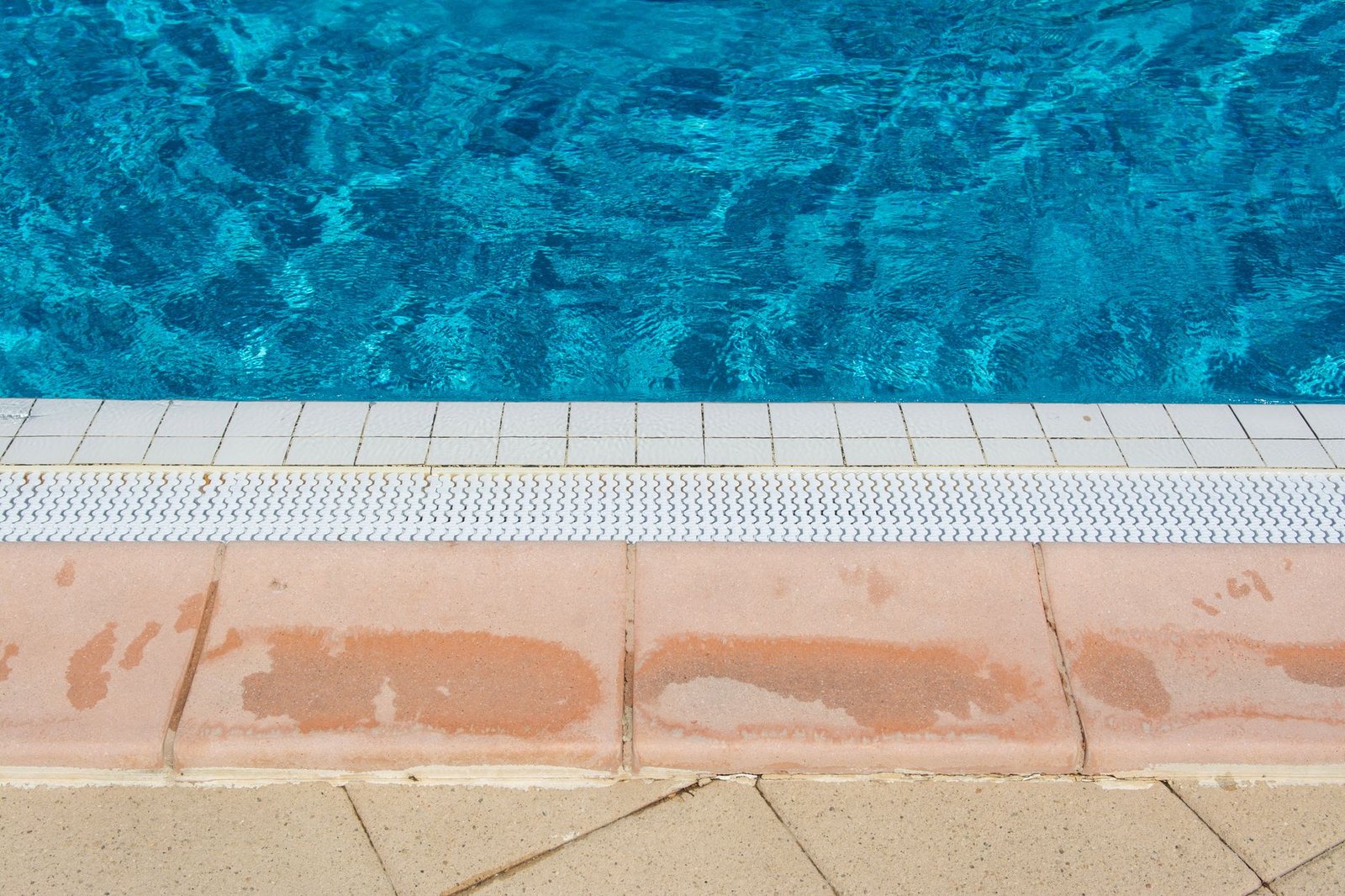 Pool edge with a row of reddish concrete coping slabs beside the overflow grate and pale deck pavers behind