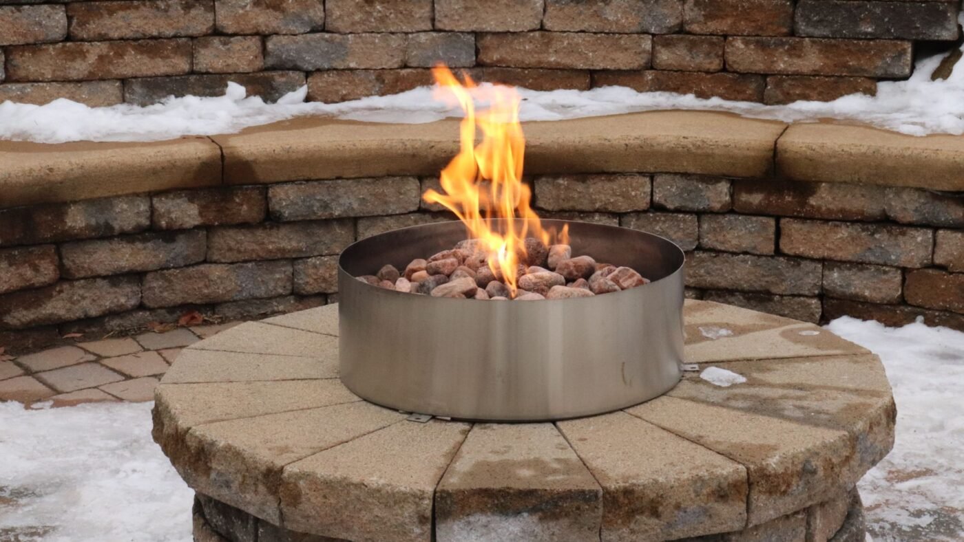 Curved capped block seat wall behind a round block fire pit with a steel insert, dusted with snow on a paver patio