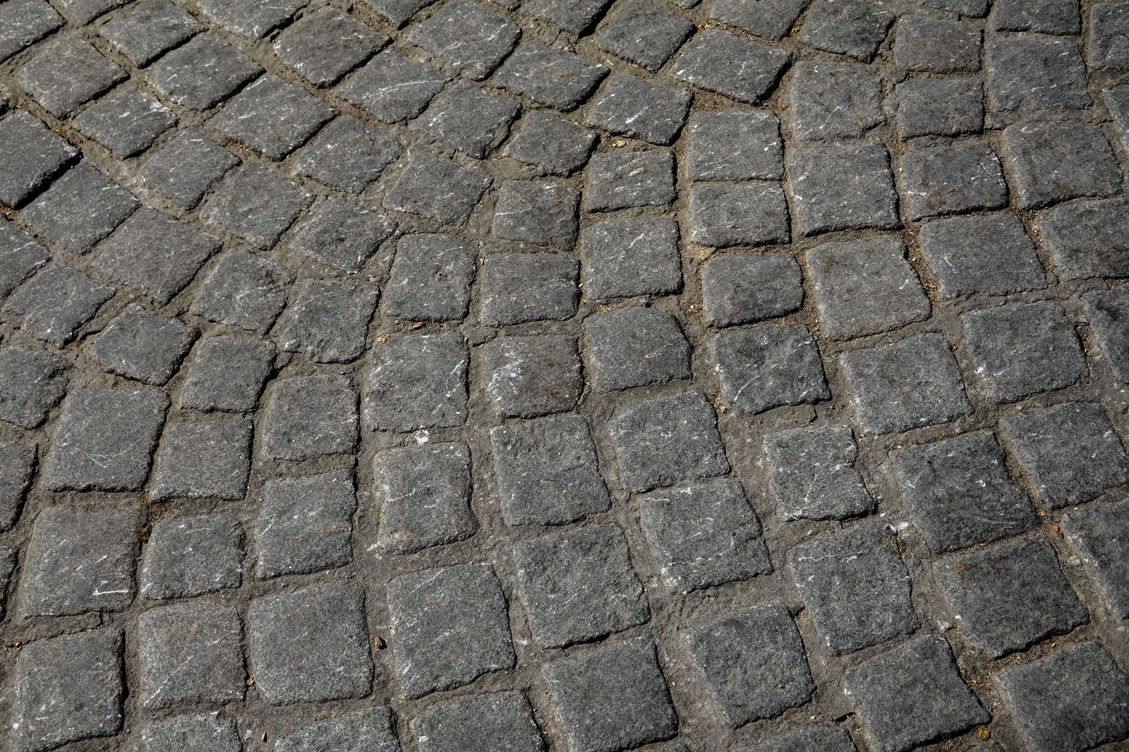 Close-up of rough-textured grey cobblestone pavers laid in an arc with sandy joints, a surface where polymeric sand residue is hard to remove